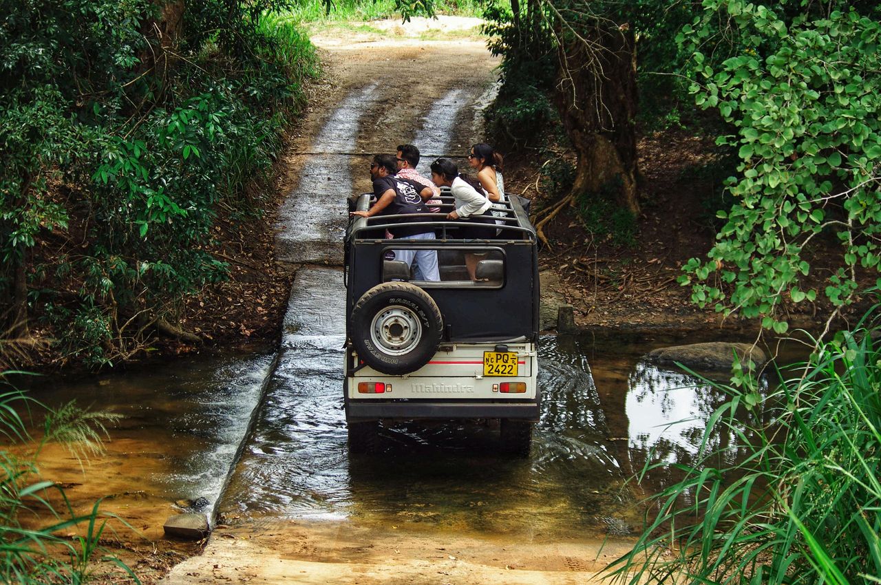 A group of adults on a jungle safari in a rugged 4x4 jeep crossing a shallow stream.